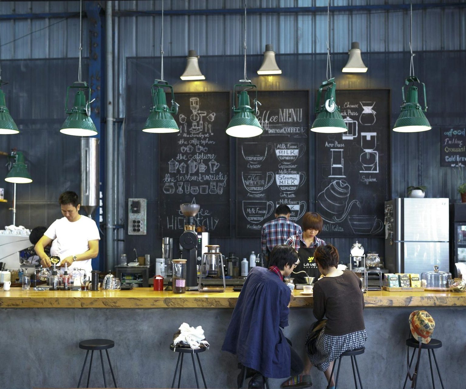 A bustling café with a modern industrial design, featuring a barista making coffee and patrons enjoying drinks.